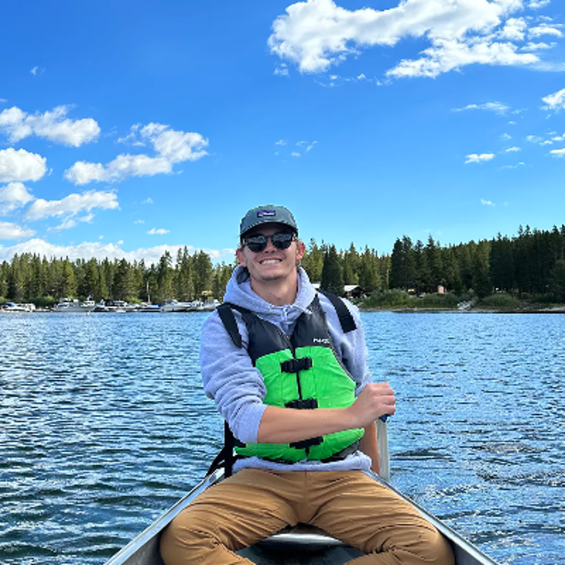 The image shows a young man in a boat on a lake. He is wearing a life vest, a hat, and a hoodie. The background features a blue sky with clouds, trees, and some buildings. The man is smiling and appears to be enjoying the day. The overall scene suggests a pleasant outdoor activity.